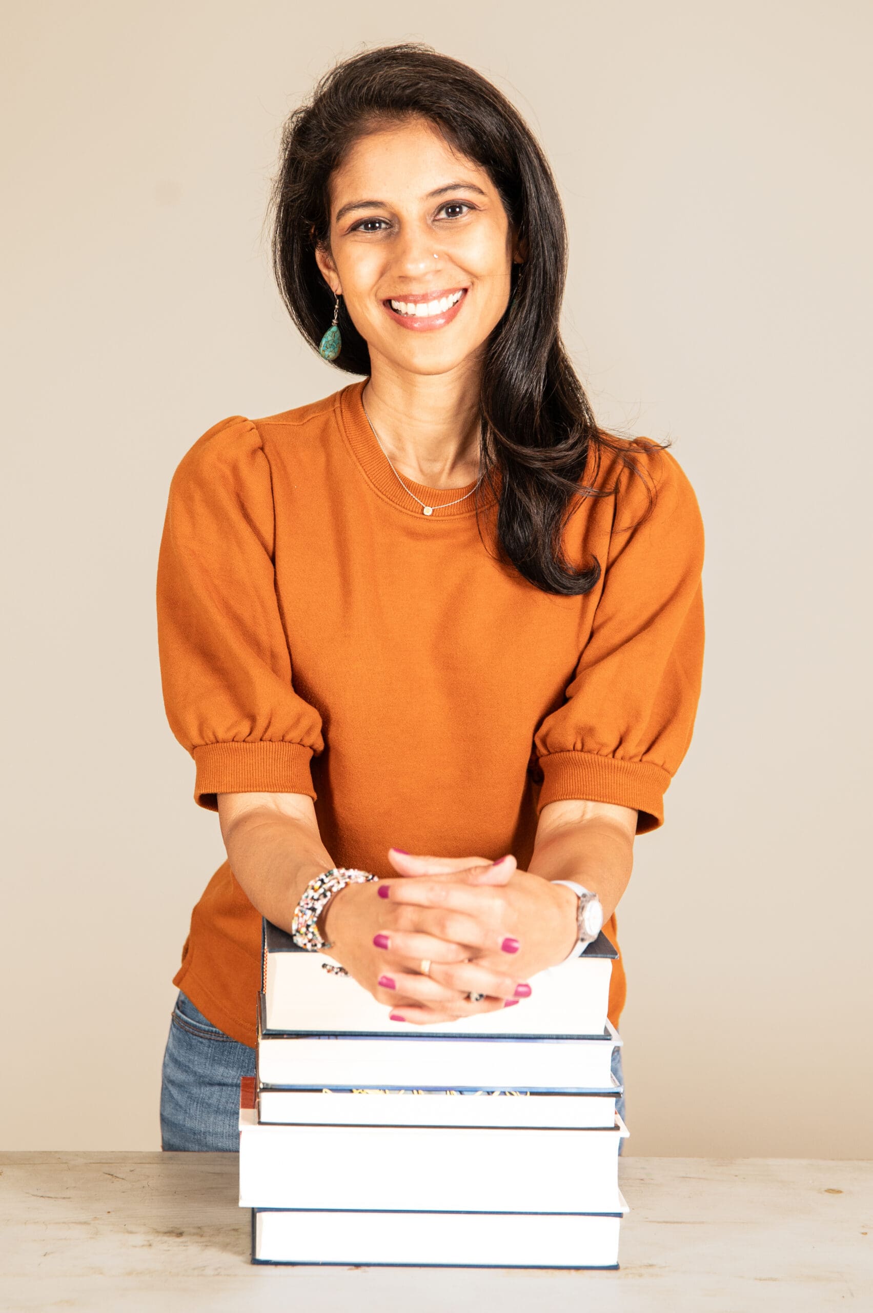 A smiling woman with long dark hair, wearing an orange shirt and jeans, leans on a stack of books.