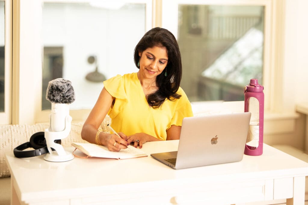 Ambitious mom in yellow blouse planning her day with notebook and laptop, creating time freedom through strategic organization.